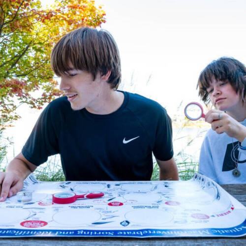Two high school students sitting a picnic table near a lake looking a sorting chart for aquatic species.